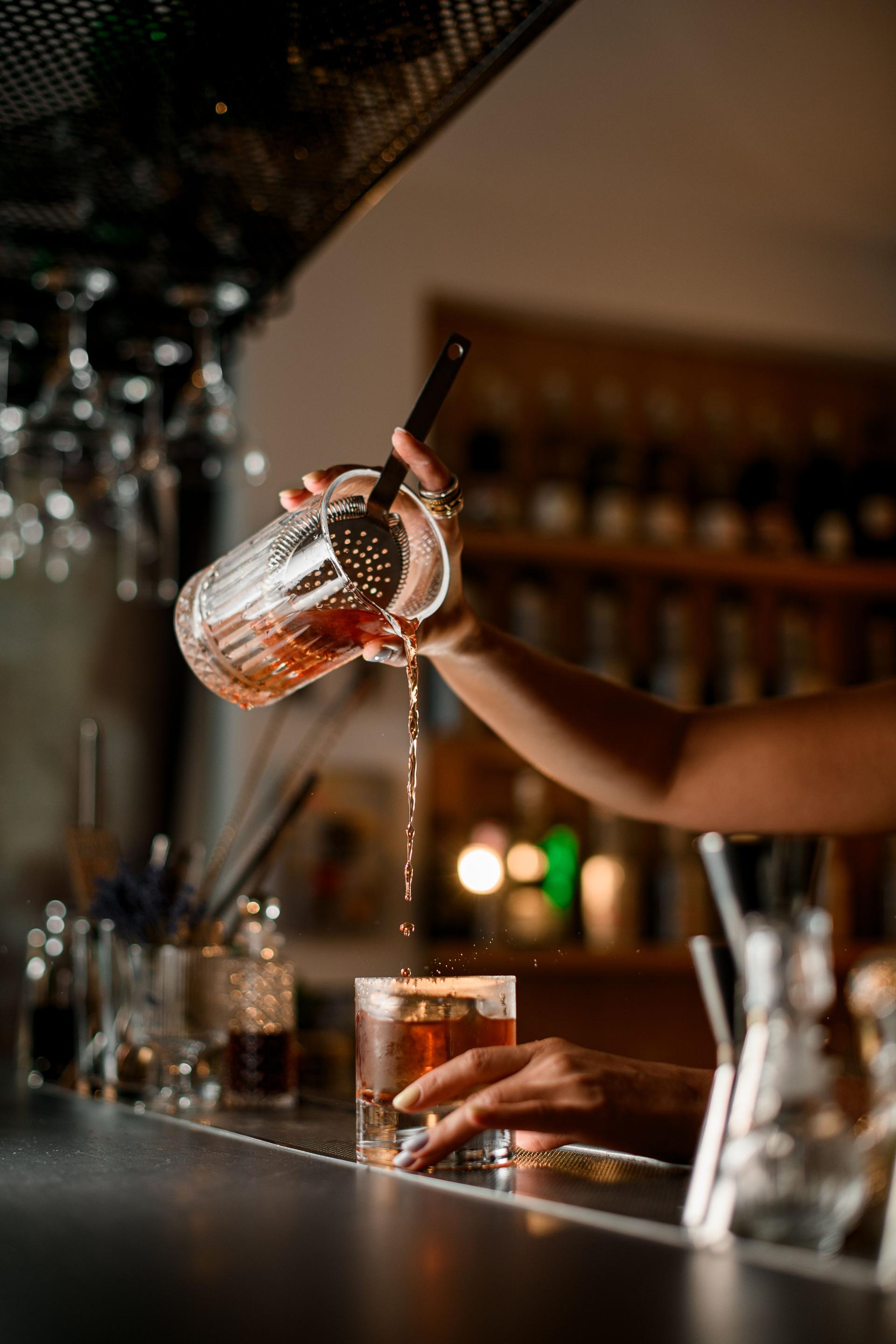 Selective focus of glass with female hands of bartender pouring cocktail from mixing glass